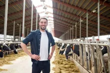 agriculture industry, farming, people and animal husbandry concept - happy smiling young man or farmer with herd of cows in cowshed on dairy farm