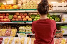 Pensive young woman looking at shelves with fresh fruits and vegetables at supermarket