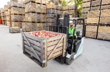 Distribution, sales, production, farm agribusiness in warehouse. Happy male driver in helmet in forklift truck lifts up red ripe apples in container on shelves, on wooden crates background, free space