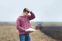 Worried young farmer standing on field and looking at papers from bank, tractor in background