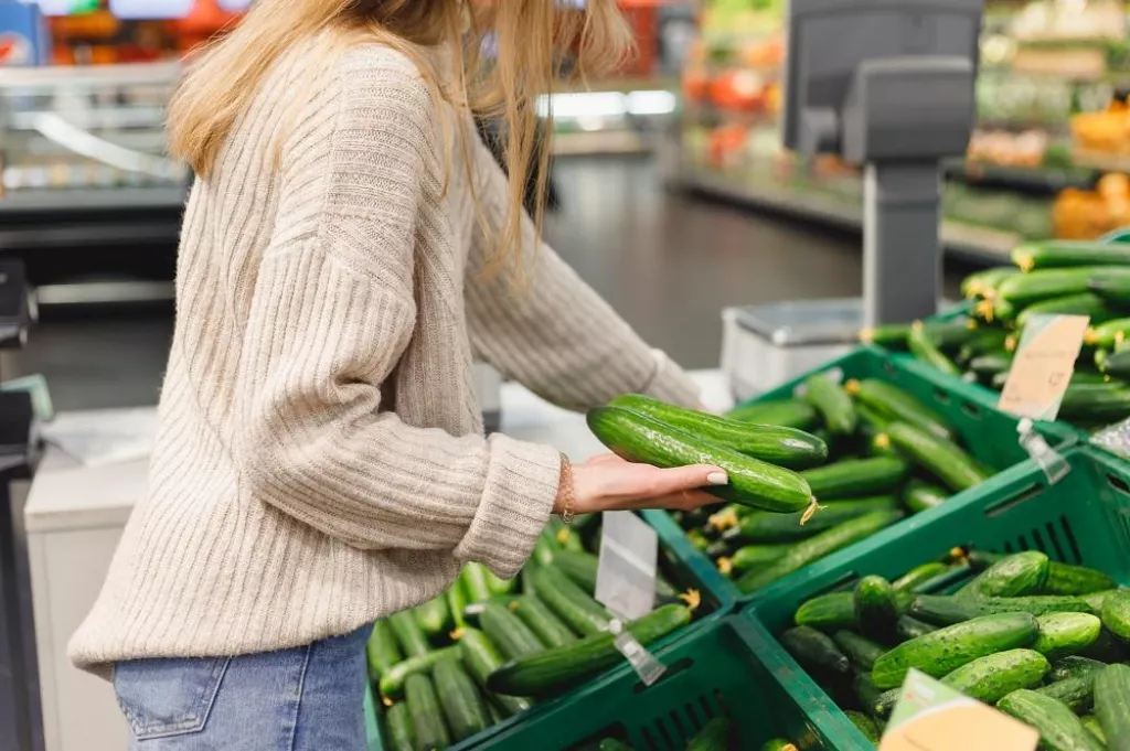 Woman pick fresh long cucumbers in shopping trolley at grocery market.