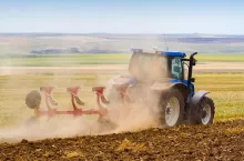 side view of blue tractor ploughing a field with trail of dust behind it
