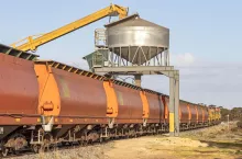 Grain train with orange freight cars (hoppers, wagons) being loaded from a cylindrical metal overhead storage bin at a remote rural location