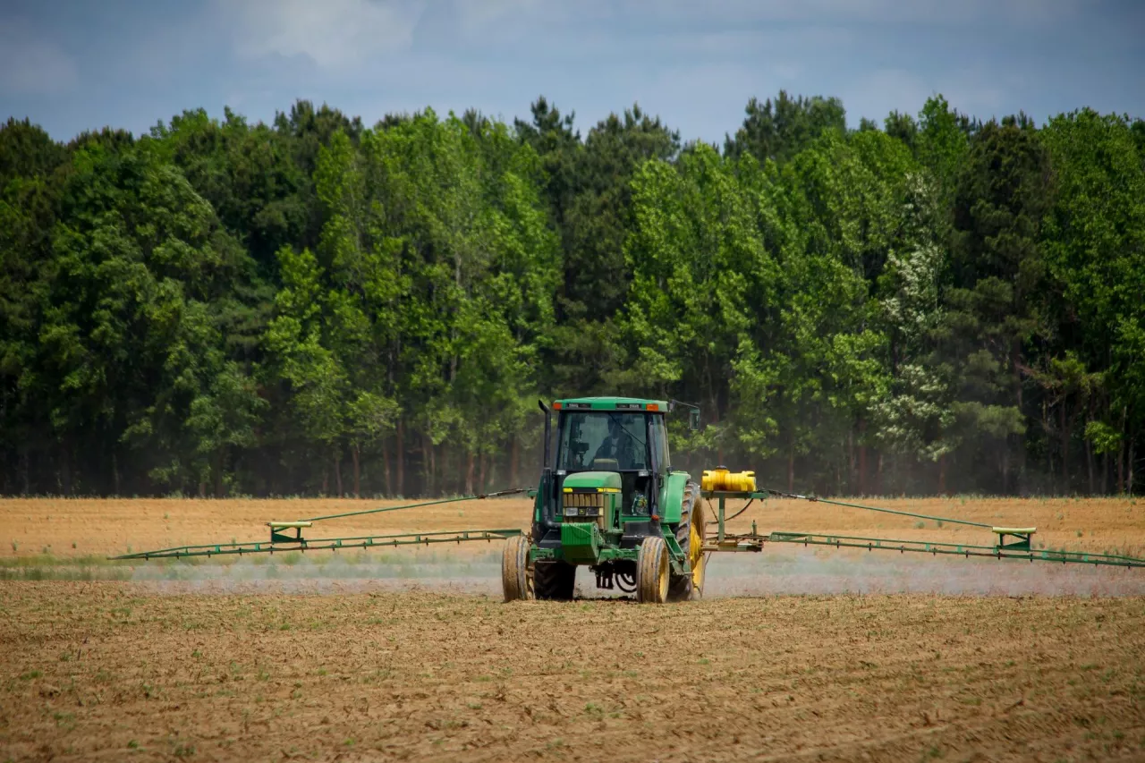 Zielony ciągnik rolniczy opryskujący pole uprawne, otoczony lasem. Maszyna wykonuje zabiegi agrotechniczne w celu ochrony upraw