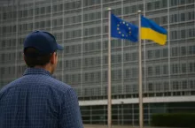 Rolnik z czapką z daszkiem patrzy w stronę budynku Komisji Europejskiej z widocznymi flagami Unii Europejskiej i Ukrainy. Obraz symbolizuje brak informacji na temat rozmów handlowych dotyczących produktów rolnych.