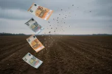Fotorealistyczne przedstawienie znikających banknotów euro nad pustym polem uprawnym. Grafika symbolizuje cięcia w budżecie Wspólnej Polityki Rolnej i mniejsze dopłaty dla rolników. Chłodny ton obrazu podkreśla powagę decyzji Komisji Europejskiej.