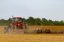 A red six wheel tractor with cabin is plowing a blank agricultural field in the spring. The attached plow tool creates a dust cloud on the farm. Image taken in Eastern Shore Maryland