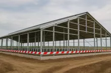 Fotorealistyczne ujęcie obory w trakcie budowy, całkowicie owiniętej czerwono-białą taśmą ostrzegawczą. Symbolizuje blokadę inwestycji rolniczych i utrudnienia w rozwoju gospodarstw.