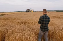 Young attractive farmer with laptop standing in wheat field with combine harvester in background