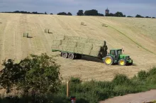Traktor ciągnie przyczepę załadowaną belami siana na zboczach pola. W tle widać wieżę zegarową.