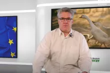 mężczyzna przed mównicą, a za nim dwa ekrany telewizyjne