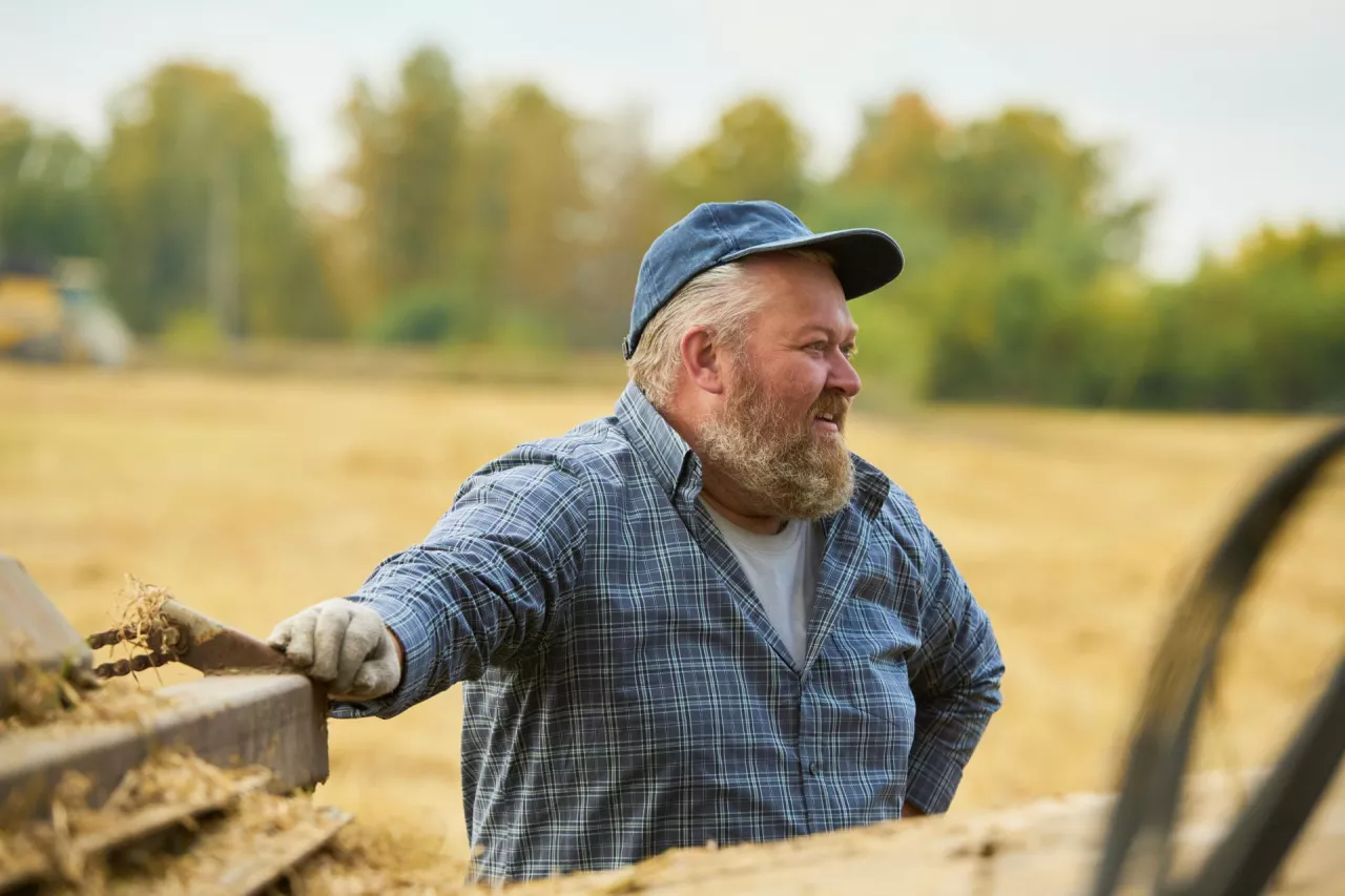 Caucasian middle aged man standing outdoors in field smiling and leaning on farming equipment, wearing cap and gloves, appearing engaged in agricultural work during harvest season