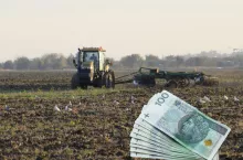 Stos banknotów 100 złotych na tle pola uprawnego po orce. W tle ciągnik z broną talerzową podczas jesiennej uprawy gleby.