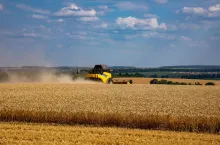 Kombajn zbożowy pracujący na polu w trakcie żniw, w tle krajobraz rolniczy i niebo z chmurami.