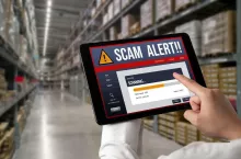 Tablet z komunikatem ostrzegawczym podczas analizy danych w magazynie logistycznym. Grafika symbolizuje cyfrowe systemy wykorzystywane do monitorowania transportów żywności i wykrywania nieprawidłowości w handlu.