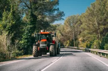 Tractor With Fertilizer Applicator With Tank In Motion On Country Road In Europe. Asphalt Road Against Background Of European Forest Landscape. Agricultural Concept