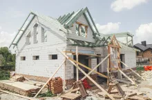Unfinished modern farmhouse building. Wooden roof framing of mansard with dormer and aerated concrete block walls with windows. Rafters and beams on blocks. New house construction